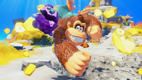 Nintendo Screenshot from Donkey Kong Bananza! shows the titular ape celebrating as fragments of a golden banana he has just uncovered explode and fill the screen around him. He grins widely as he jumps in the air, clenched fist outstretched towards the viewer. Next to him, a small, purple rock with blue gem eyes also leaps for joy.