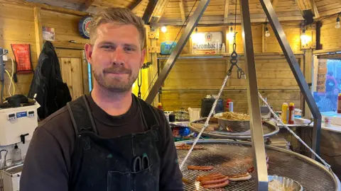 A man standing in a wooden hut with two large circular barbecues with sausages on top. He is wearing a brown top with a black apron over the top.