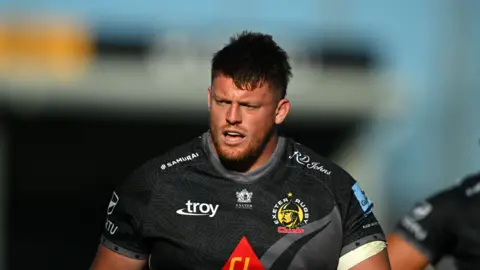 Will Goodrick-Clarke wearing a black Exeter Chiefs jersey. He has short dark hair and a gingery beard. 