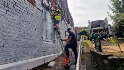 BBC/Naj Modak Zoe Genders wearing a high-visibility vest stands on a ladder painting over graffiti on the white-painted brick wall of a red-brick building. Another person steadies the ladder from the ground. A white work van with its rear doors open is parked nearby, with ladders and painting equipment visible around the site. The scene is outdoors on a narrow pathway beside the building under an overcast sky.