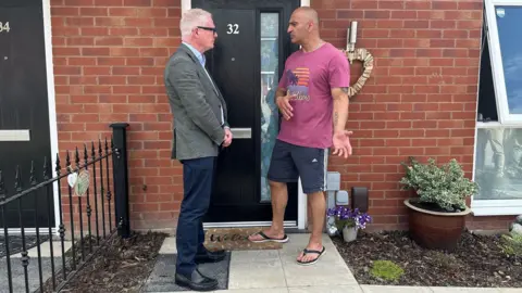 Man in a green suit jacket standing on a doorstep talking to a man in a t-shirt, flipflops, and shorts