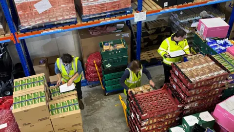 The image shows the inside of a large warehouse where food is being sorted and organised. There are tall metal shelving units stacked with pallets of packaged goods, including boxes and shrink-wrapped items. On the floor, there are crates of fresh produce, such as a wooden box filled with green apples, and stacks of packaged bread rolls. Three people wearing high-visibility vests are working among the crates and boxes.