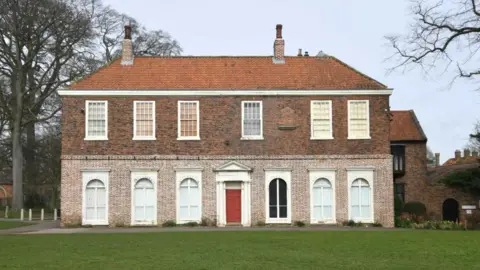 Donna Clifford A large brick-built Georgian building, with a red tile roof and red front door. There is a lawn to the front of the building.