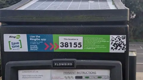 BBC/Leigh Jones A close-up shot of a solar-panel powered pay and display machine. It's made of black metal and stuck on is a professionally printed RingGo branded information sticker denoting the location code for the car park the machine is located. On the right is a pixelated QR code stuck conveniently over a white box in the graphic allowing it to seamlessly fit with the branding. 