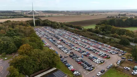 SHAUN WHITMORE/BBC An aerial view of the Queen Elizabeth Hospital's car park, where the new hospital will be built. It shows rows and rows of cars in a large car park surrounded by trees and fields.