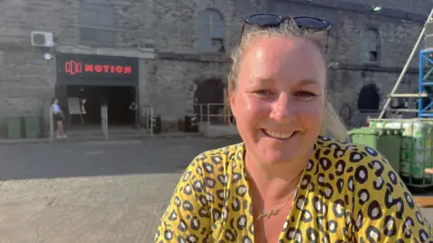 Carly Heath in a yellow leopard print outfit and gold necklace that says Junglist. She is sitting at a table outside Motion. You can see the main entrance behind her with the red Motion logo above it.