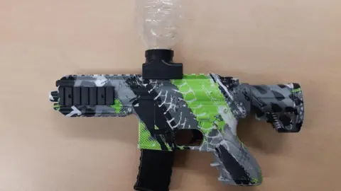 A gel blaster imitation gun on a wooden table top. The imitation firearm is patterned in grey, black and green army camouflage print, with a see-through plastic bottle attached at the top.