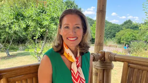 Charlotte Rose/BBC A woman wearing a green dress with a white, orange and yellow scarf around her neck.
She is smiling while sitting on the bench in the garden.