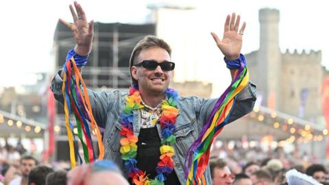Man with sunglasses on and a rainbow garland around his neck and rainbow streamers on his wrists