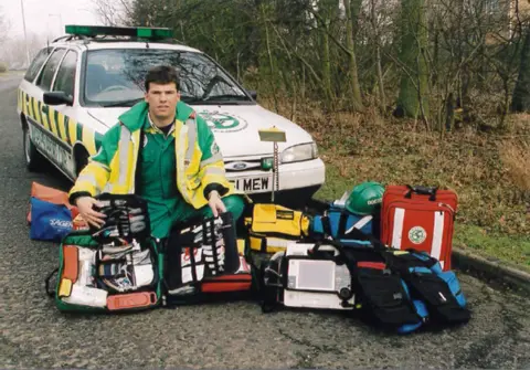 Magpas Dr Rod Mackenzie in the early 2000s. He is kneeling on the ground with various medical kit in front of him, while behind him is a car in Magpas livery. He is wearing a green and yellow coat over a green jumpsuit and has short brown hair. 