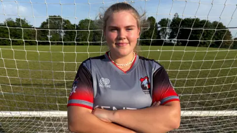 A woman with arms folded, smiling towards the camera, stood in the middle of a football net, wearing a Ravenshead Red shirt.