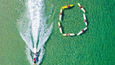 An overhead shot shows a group of people in canoes on the water forming a circle as a boat passes them