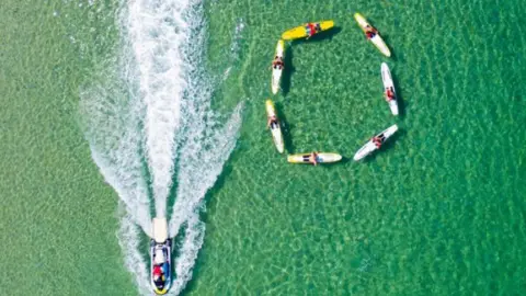 RNLI An overhead shot shows a group of people in canoes on the water forming a circle as a boat passes them