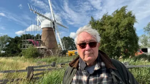 Andrew Turner/BBC Adam Singer is wearing a checked shirt and wax jacket, and sun glasses. He has white hair swept over to the right. He is standing near the Toft Monks Wind Pump, with reeds and trees nearby, and the pump bearing its sails.