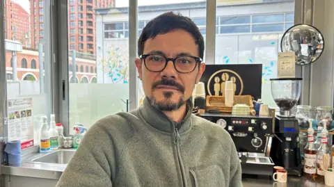 A man wearing glasses and a grey zip-up fleece standing behind a coffee machine in a café setting, with cups, bottles, and a window overlooking city buildings in the background