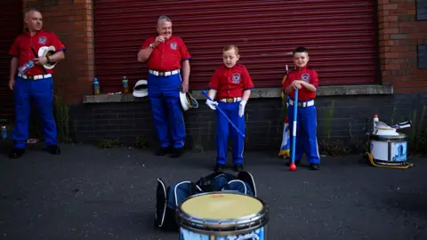 PA Media Members of the orange Order parades wait on Clifton Street dressed in red tops, blue trousers and surrounded by drums. 