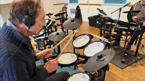 Richard Alcock is sitting at an electric drum kit. He is wearing a blue jumper. He is sitting opposite another person playing an electric drum kit.