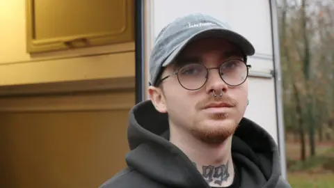 A man with a grey/blue baseball cap and large round glasses stands in front of a caravan. The door is open and a yellow glow emanates from inside. He's got a septum piercing - a ball on each end dangles from his nose - and a tattoo reading "1991" in ornate gothic script on his lower neck. He wears a dark coloured hoodie.