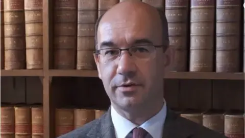 West Northamptonshire Council Man with short dark hair and glasses stands in front of leather-bound books