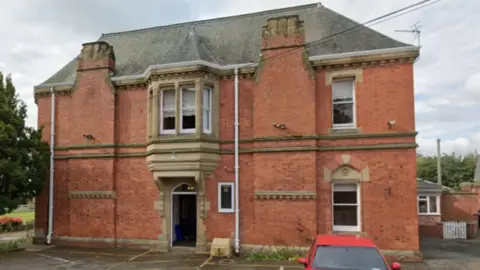 A two-storey red brick building with a sloped grey roof