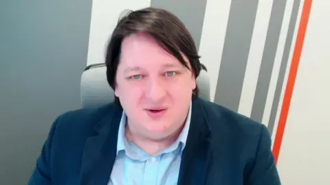 A man looks at the camera. He wears a navy jacket and blue shirt. He has dark mid length hair and blue eyes.