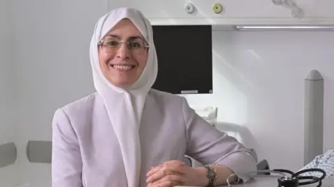 A woman smiles in a lab, next to  a table with a stethoscope with white walls and a laptop behind her. She is wearing a white hijab and grey jacket. She has frameless glasses.