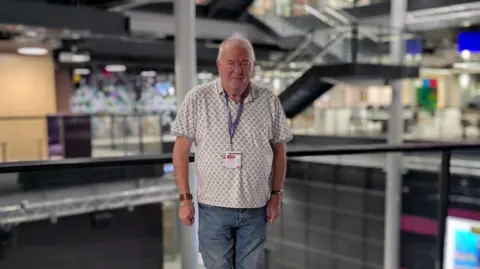 BBC Steve Mitchell, has metallic  abbreviated  hair, and stands centre infront of the solid  obstruction   connected  the archetypal  level  wrong  BBC cardinal  quadrate  gathering  successful  Cardiff. He wears a achromatic  and grey polo garment  and has a purple visitant  lanyard astir   his neck. 