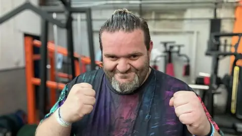 Ben Bretts, a man wearing a black t-shirt with a multi-coloured pattern on it, is holding his hands up in fists, mimicking a boxing pose.