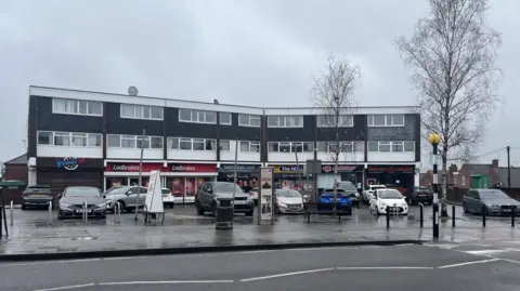 The Hollies in Eastwood is pictured. It is a three-storey building with shops on the bottom floor. Cars are parking in the car park.