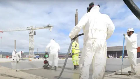 Workers wearing white suits and florescent overalls lay concrete at the treatment plant in Sellafield. There is a crane in the distance and other industrial buildings.
