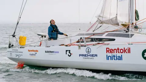 Premier Marinas Mr Ketchell is sailing aboard Mindset. He is wearing the same outfit, a blue waterproof long sleeved jacket with a black buoyant vest on. He is in the middle of the ocean and his yacht is while with various business sponsor logos on the exterior.