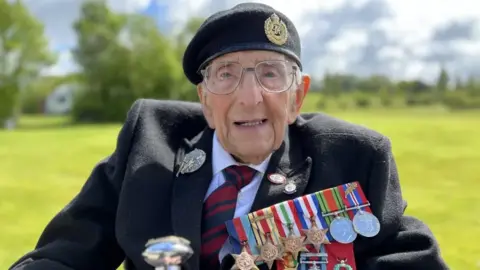 Don Sheppard, aged 103, smartly dressed in a black coat and adorned with medals. He is outside with grass and trees behind him.