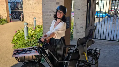 Layla Moran, wearing a cycle helmet, stood holding up her cargo bike with a child seat on the back.