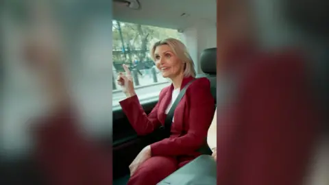 A woman in a red trouser suit sits in a car and points at something out the window