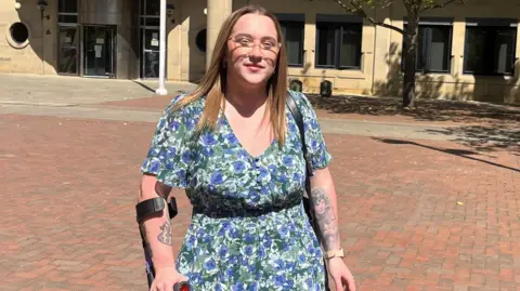 A woman in her 20s holding a crutch wearing a flowery dress outside a court building. She has tattoos on her arms, is wearing glasses and has light brown coloured hair. 