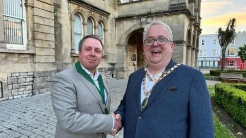 North Somerset Council A man on the right in a grey suit jacket and a green ribbon with a medal on it. He is shaking hands with a man on the right who is wearing ceremonial chains and a navy suit jacket. Both men wear white shirts and smile at the camera. Behind them is a stone building.