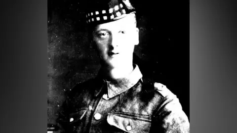 A grainy, black-and-white photo of a pictured in military fatigues. He is wearing a military-style cap. The light is striking the side of his face which appears in high contrast against a dark background.
