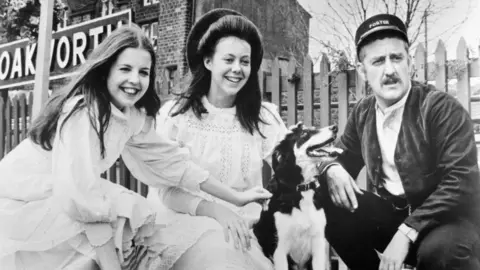 Getty Images A black and white photo of actors Sally Thomsett and Jenny Agutter meeting Bernard Cribbins in a scene from the film The Railway Children. All three are crouched down on a railway station platform next to a dog.
