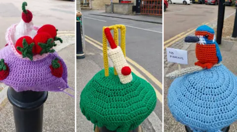 Crocheted yarn covering on a black bollard decorated with red strawberries and a swirl of white cream, resembling a dessert, a green crocheted cover on a bollard topped with miniature knitted cricket stumps, bat and ball, and a small crocheted bird in blue and orange perched on a bollard cover holding a sign reading “No Fishing”, with parked cars in the background, all by a road. 