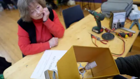 A woman in a red top and a black cardigan watches as her lamp in a mustard yellow lampshade is repaired.