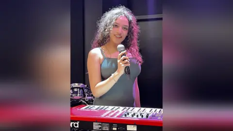BBC Iquao Aluko in a music studio at the college holding a microphone. She has curly hair and wears a blue dress and hoop earrings.