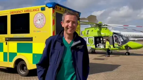 Michael Griksaitis standing by an ambulance and helicopter. He is smiling and wearing a blue jacket and a green top. There is a person standing next to the green ambulance wearing a florescent jacket. Both venicles are on a brown paved area with clouds in the sky above.