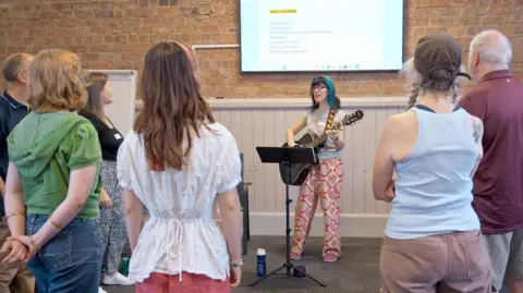 A group of people stand together, looking at a woman playing a guitar. 