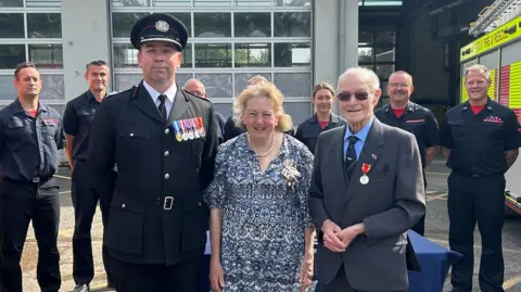 Norfolk Fire and Rescue Service Three people are standing in the forefront of the image. Two men are on the ends wearing suits with medals on their jacket, and a woman is standing in the middle wearing a blue dress. People in uniform are standing in the background.