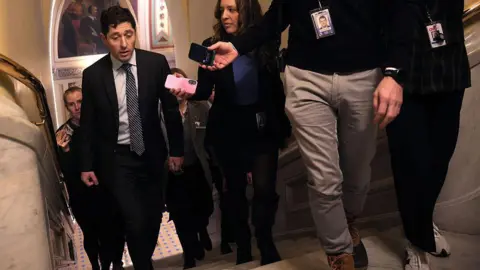 Getty Images Frey in dark suit and black-and-white tie walks up stairs surrounded by reporters holding their phones out to record him