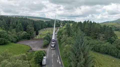 CWP Energy A turbine blade lifter passing through southern Scotland on a road with trees at its side, what looks like a graveyard on the left and an operational wind farm in the distance.