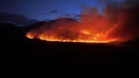 A large wildfire on a mountain at night. Smoke is bellowing into the sky. The rest of the mountain is in darkness. The sky is dark navy with some clouds. 