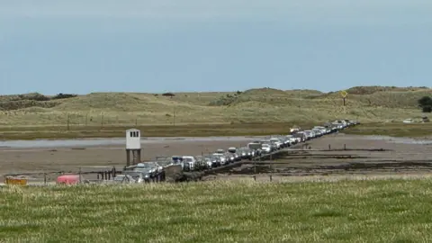 James Gillespie A queue of cars on a road surrounded by sand. Sand dunes can be seen in the background.