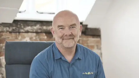 Sue Lambert Trust Clive Evans is sitting in a chair with a sky-light window behind him. He's wearing a blue shirt.