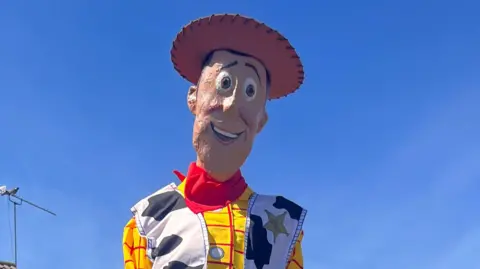 A life-sized model of Woody from the animated movie Toy Story. He is wearing a brown cowboy hat, yellow and red chequered shirt, a jacket with a cow print on it and blue trousers. 